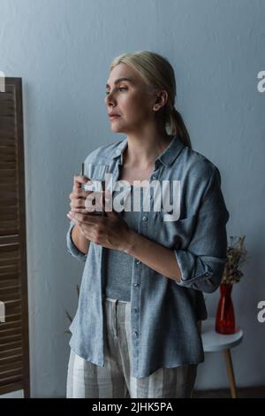 Blonde Frau mit Wechseljahren hält ein Glas Wasser und schaut weg Stockfoto