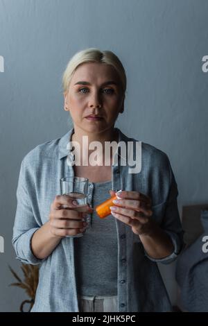 Blonde Frau mit Wechseljahren hält ein Glas Wasser und eine Flasche mit Medikamenten Stockfoto