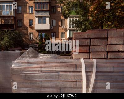 Ein Stapel roter Pflasterfliesen im Vordergrund und ein altes rotes Ziegelgebäude, das im Hintergrund von untergehenden Sonnenstrahlen in Kiew, Ukraine, beleuchtet wird. Straße im Bau Stockfoto