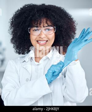 Porträt einer jungen afroamerikanischen Wissenschaftlerin mit afrohaarigen Haaren, die einen Laborkittel und eine Brille trägt, während sie im Labor Handschuhe anlegt. Gemischt Stockfoto