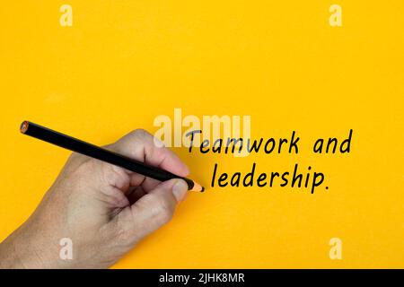 Männliche Hand schreibt in schwarzem Bleistift das Wort TEAMWORK UND FÜHRUNG auf einem gelben Hintergrund. Ermutigende Worte und Geschäftskonzept. Stockfoto