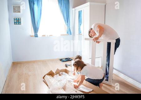 Papa, kleine Mädchen Töchter, die nach den Anweisungen im Kinderzimmer ein hölzernes Babybett zusammenstellen. Vater helfen, Kindermöbel montieren. Selbstbedienung Stockfoto