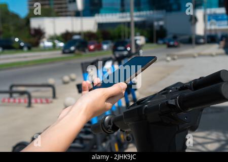 Nahaufnahme die Hand einer Frau scannt mit einem Mobiltelefon den QR-Code eines Rollers in der Stadt. Städtischer Fahrstil auf ökologischen Verkehr. Stockfoto