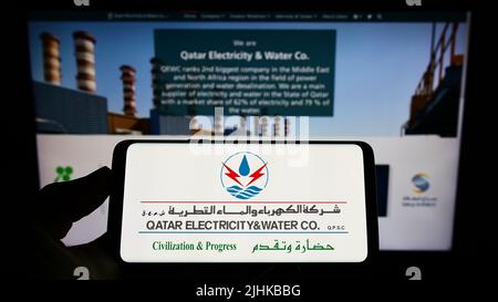 Person, die Mobiltelefon mit Logo der Firma Qatar Electricity Water Company (QEWC) auf dem Bildschirm vor der Webseite hält. Konzentrieren Sie sich auf die Telefonanzeige. Stockfoto