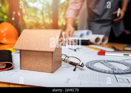 Nahaufnahme eines Hausmodells auf dem Schreibtisch mit Architekten, die mit Bauplan-Bauprojekt, Architekturkonzept, arbeiten. Stockfoto
