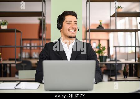 Lächelnder junger hispanischer Verkäufer mit Headset hält einen Online-Anruf ab, ein männlicher Kundendienstmitarbeiter sitzt vor einem Laptop und schaut weg Stockfoto