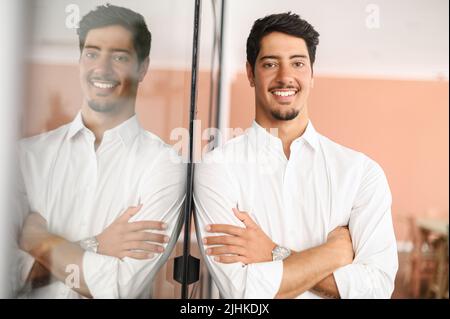 Lächelnder hispanischer männlicher Mitarbeiter in einem eleganten, lässigen Hemd steht mit gekreuzten Armen im modernen Coworking Space, ein fröhlicher, stolzer lateinamerikanischer Geschäftsmann blickt auf die Kamera, die drinnen posiert Stockfoto