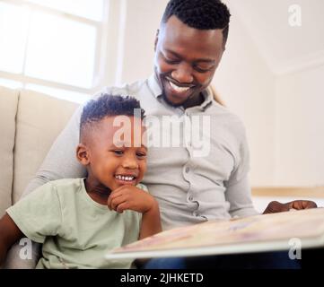 afroamerikanischer Vater, der sich zu Hause mit seinem Vater verbunden hat. Schwarzer Mann hilft seinem Sohn ein Buch zu lesen und zu lernen, während er zu Hause auf einem Sofa sitzt Stockfoto