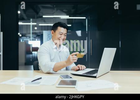 Glücklicher asiatischer Geschäftsmann, der in einem modernen Büro arbeitet, Mann, der Online-Geldtransfer macht, einen Laptop verwendet und eine Bankkreditkarte hält Stockfoto