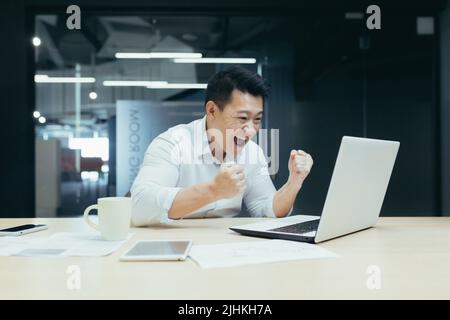 Porträt des erfolgreichen asiatischen Chefs, Geschäftsmann, der auf den Laptop-Bildschirm schaut, gute Nachrichten liest und Sieg und Erfolg feiert, Mann bei der Arbeit Stockfoto