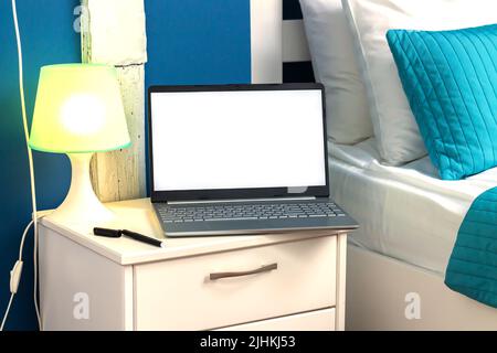 Offener Laptop-Computer mit leerem weißen Bildschirm neben der Lampe auf dem Nachttisch Stockfoto