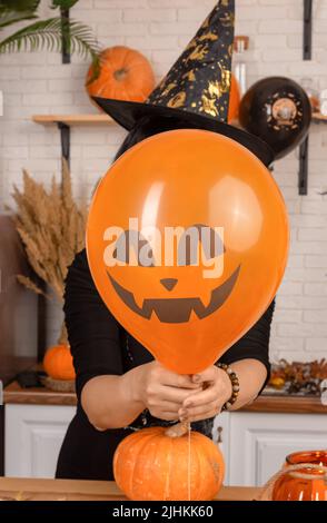 Eine unkenntliche Frau, die ein gruseliges Halloween-Gesicht vor ihrem Gesicht hält, einen leuchtend orangefarbenen Ballon. Eine Frau in einem spitzen Karnevalshut verbirgt ihr Gesicht Stockfoto