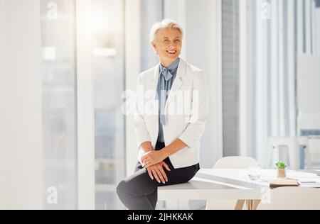 Porträt einer selbstbewussten reifen kaukasischen Geschäftsfrau, die allein in einem Büro auf einem Tisch sitzt. Nur eine Frau mit grauem Haar lächelnd und aussehend Stockfoto