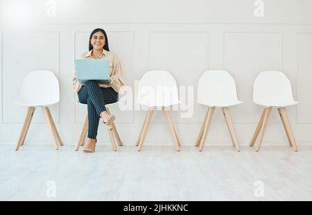 Junge glücklich lächelnde Mischling hispanische Geschäftsfrau, die am Laptop arbeitet, während sie auf ein Interview wartet, das auf einem Stuhl gegen eine weiße Wand in einem sitzt Stockfoto