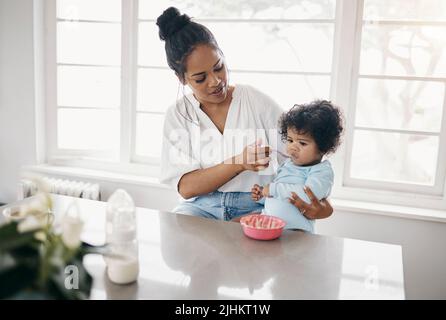 Sie müssen essen. Schoss eine attraktive junge Frau, die mit ihrer Tochter in der Küche saß und sie fütterte. Stockfoto