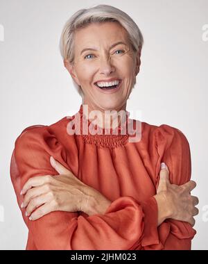 Fröhliche, reife Frau, die lacht und sich vor einem grauen Studiohintergrund umarmt. Glückliche ältere Frau, die sich umarmt und liebt. Selbstbewusst Stockfoto