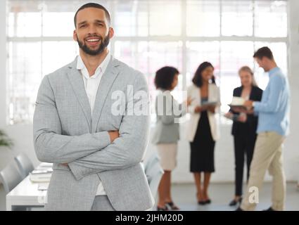 Junger glücklicher gemischter Geschäftsmann, der mit gekreuzten Armen in einem Büro mit Kollegen steht. Hispanischer männlicher Chef bei einem Treffen mit Kollegen Stockfoto