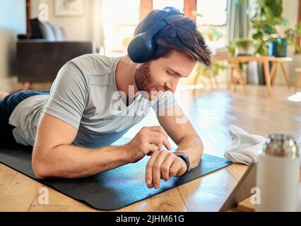 Ein junger Kaukasusmann hörte Musik mit Kopfhörern und überprüfte die Zeit auf einer digitalen Armbanduhr, während er zu Hause trainiere. Typ, der Fitness trägt Stockfoto