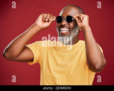Glücklicher reifer afroamerikanischer Mann, der allein vor einem roten Hintergrund in einem Studio steht und mit einer Sonnenbrille posiert. Lächelnder schwarzer Mann Stockfoto