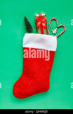 Weihnachtsstrumpf mit kleiner Geschenkbox, Bäumen und Zuckerrüben in Herzform auf grünem Hintergrund mit Platz für Text. Flache Lay-Zusammensetzung von Hol Stockfoto