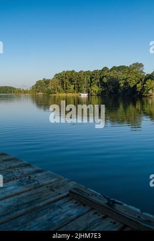 Ein hölzerner Dock mit einem Stutzen an einer Bucht in der Nähe des Atlantischen Ozeans mit Blick auf ein Ufer mit Bäumen und ein Segelboot in der Ferne angedockt. Stockfoto