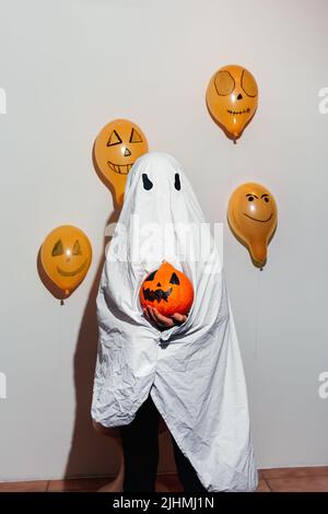 Frohe Halloween. Der Geist hält den orangefarbenen Kürbis in der Hand. Kind in weißem Anzug, bemalte gelbe Luftballons. Verschiedene Emotionen von Freude, Wut, Lachen. Festliches Design, Partykonzept. Weicher selektiver Fokus Stockfoto