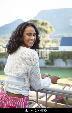 Biracial junge Frau mit lockigen Haaren stehend am Geländer im Balkon gegen Berg und klaren Himmel Stockfoto