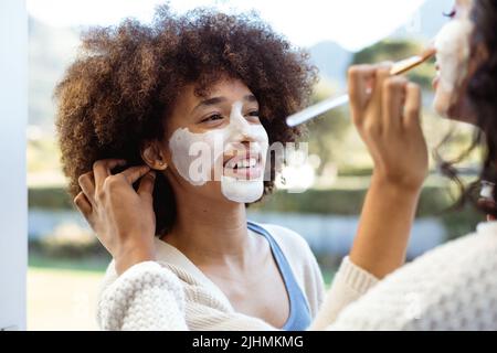 Lächelnde Biracial Freundinnen, die Schönheitscreme auf die anderen Gesichter auf dem Balkon auftragen Stockfoto