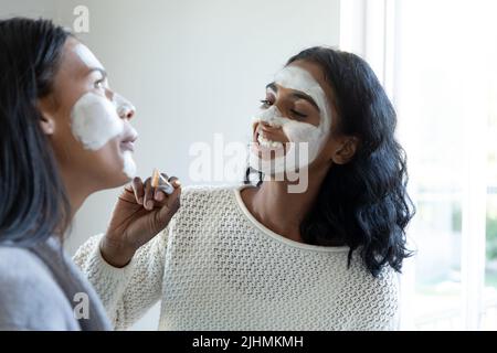 Biracial lächelndes junges Weibchen, das Gesichtscreme auf das Gesicht eines tausendjährigen Freundes zu Hause anwendet Stockfoto