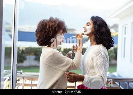 Seitenansicht von biracial weiblichen Freundinnen, die Schönheitscreme auf die anderen Gesichter im Balkon auftragen Stockfoto