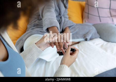 Mittelteil einer jungen Birazialfrau, die die Fingernägel einer Freundin malt, während sie zu Hause auf dem Bett sitzt Stockfoto