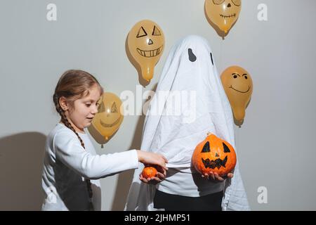 Frohe Halloween. Mädchen gibt dem Geist einen orangefarbenen Kürbis. Kind im weißen Anzug. Lackierte gelbe Luftballons. Verschiedene Emotionen von Freude, Wut, Lachen. Festliches Design, Partykonzept. Weicher, selektiver Fokus Stockfoto