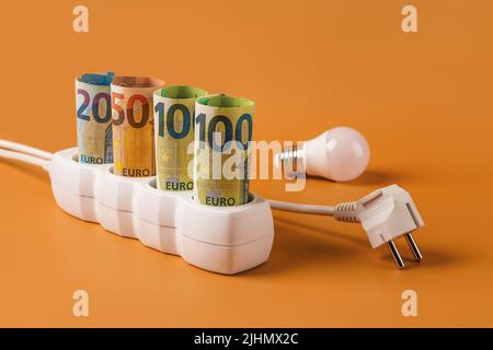 Konzept der steigenden Energiekosten. Euro-Geldscheine, die in eine weiße Steckerleiste, einen elektrischen Stecker und eine Glühbirne auf orangefarbenem Hintergrund eingesteckt sind. Energiekosten. Stockfoto