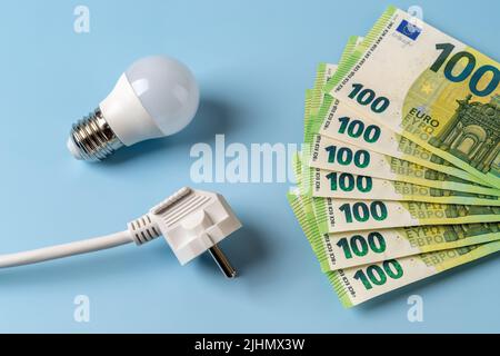Anstieg der Strompreise. Weißer elektrischer Stecker, Glühbirne und 100-Euro-Banknoten in Fächerform auf blauem Hintergrund. Steigende Stromrechnung. Stockfoto