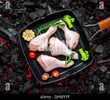 Rohe Hähnchenbeine in einer Pfanne mit Gemüse und Gewürzen stehen auf einem heißen Barbecue-Grill Stockfoto