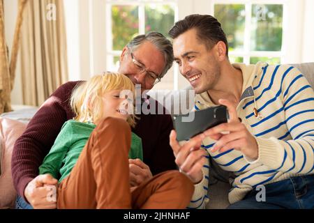 Bild von glücklichen kaukasischen Männern der drei Generationen, die auf dem Sofa sitzen und das Smartphone benutzen Stockfoto