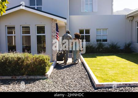 Rückansicht eines mehrrassischen Soldaten in Tarnung, der die Hand seiner Frau hielt und in das Haus ging Stockfoto