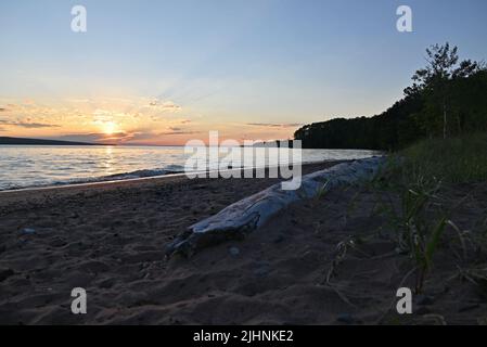 Ein gefallener Baum am Strand, während die Sonne am Lake Superior bei Sonnenuntergang auf Oak Island im Apostle Island National Lakeshore reflektiert. Stockfoto