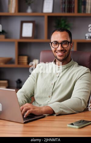 Porträt eines jungen afrikanisch-amerikanischen Geschäftsmannes, der im Büro mit einem Laptop arbeitet. Lächelnder selbstbewusster Unternehmer, der Brillen mit Computer trägt Stockfoto
