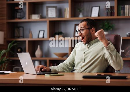 Glücklicher afroamerikanischer Geschäftsmann, überrascht von guten Nachrichten und einer Ja-Geste, während er auf einen Laptop im Büro schaute. Der Manager genießt den Erfolg Stockfoto