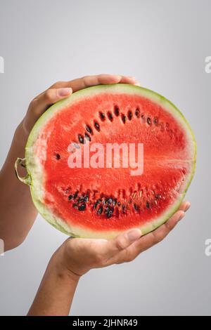 Die Hälfte der Wassermelone in den Händen einer unbekannten kaukasischen Frau, die sie vor dem Hintergrund der weißen Wand hält Stockfoto