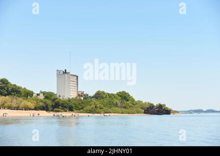 Dec 30, 2021, Colon, Entre Rios, Argentinien: Strand von einem Boot auf dem Uruguay-Fluss aus gesehen, Leute, die das Ufer genießen, Intersur-Hotel und verlassene ru Stockfoto