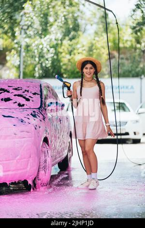 Frau mit Schlauch steht mit dem Auto in rosa Schaum bedeckt Stockfoto