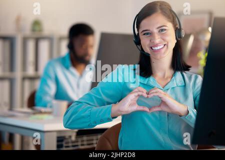 Porträt einer glücklichen jungen lächelnden kaukasischen Callcenter-Telefonverkäuferin, die mit ihren Händen die Herzform streicht, während sie im Büro am Headset spricht Stockfoto