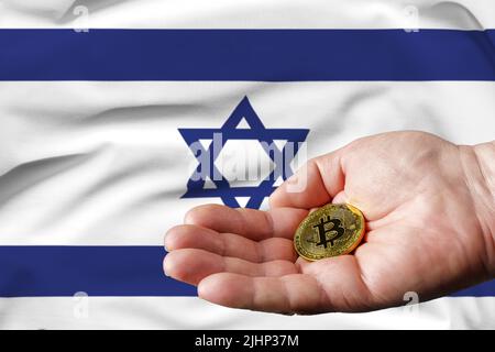 Goldene Bitcoin-Münze in der Hand des Menschen, Israel-Flagge im Hintergrund. Stockfoto