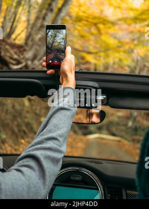 Der Mensch nimmt ein vertikales Video auf einem Mobiltelefon vom Auto auf der Straße im herbstlich schönen Wald Stockfoto
