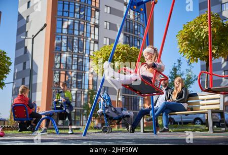 Glückliche Familie - Vater, Mutter und Kinder, die sich am sonnigen Morgen gemeinsam amüsieren. Eltern sitzen auf der Bank, während Kinder auf dem Spielplatz spielen. Moderne Wohngebäude im Hintergrund. Stockfoto