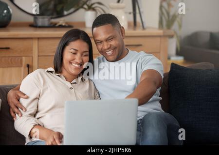 Attraktives junges gemischtes Paar, das sich auf dem Sofa entspannt und einen Laptop benutzt. Lächelndes afroamerikanisches Paar, das sich Filme ansieht, während es liebevoll ist Stockfoto