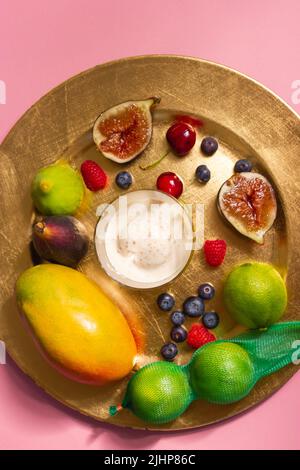 Ein goldener Teller, eine Platte mit frischen tropischen Früchten und ein Eisjoghurt auf einer rosafarbenen Tischdecke auf einem Tisch. Feigen, Mango, grüne Limetten, Kirschen und würzen Stockfoto
