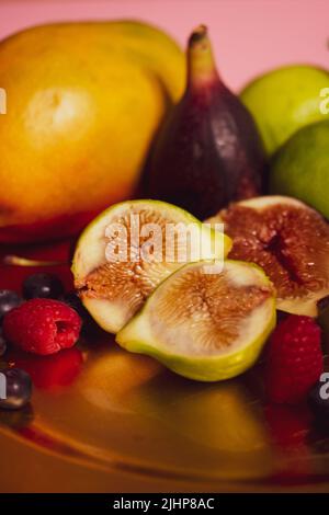 Auf einem Tisch steht ein goldener Teller mit frischen tropischen Früchten. Halbiert geschnittene Feigen, Mango, Limetten und saisonale blaue Beeren, Himbeeren. Vielzahl von s Stockfoto